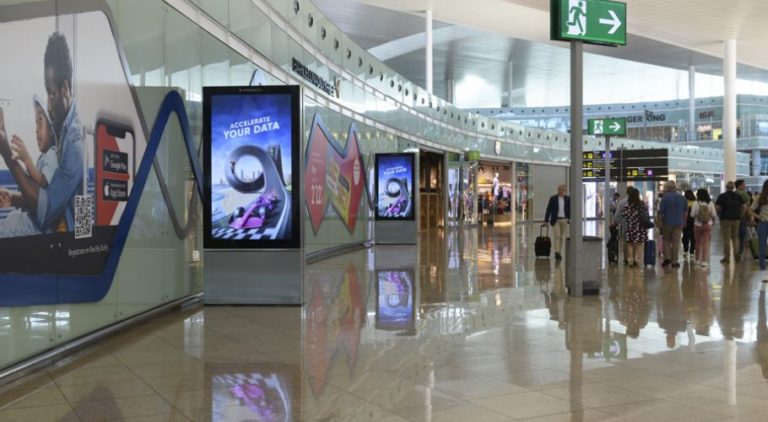 On the Barcelona Airport, on the right side of the image, several people are visible, and on the left in the foreground, two digital City Light Posters are standing in a row, displaying advertising for Ataccama Software.