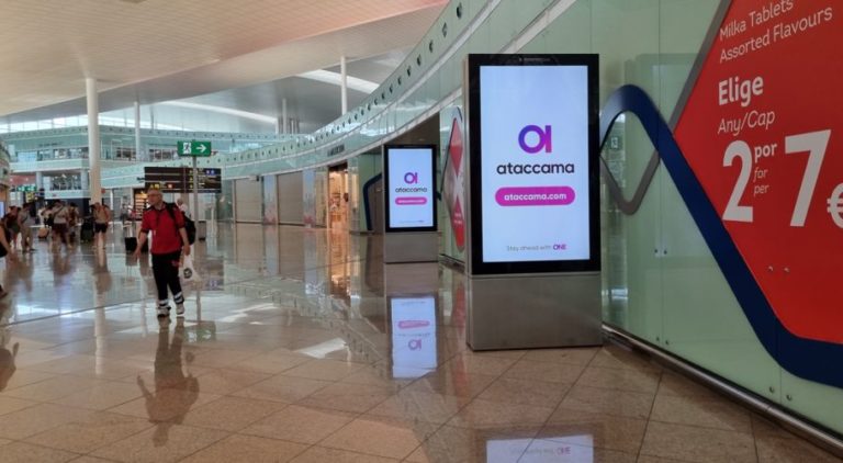 The image shows people passing by on the left at Barcelona Airport, with two digital City Light Posters in the foreground. The digital screen displays advertising for Ataccama Software.
