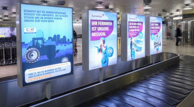 The lightbox is located at the arrivals area in Hannover Airport. People are waiting at the baggage claim. The billboard advertisement showcases the advertisement for CEAT.