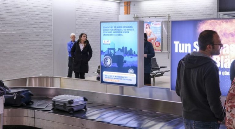 At Hannover Airport at the arrivals area, on the right side of the image, a lightbox is visible. Next to it, people are waiting for their luggage. The billboard advertisement displays an advertisement for CEAT.
