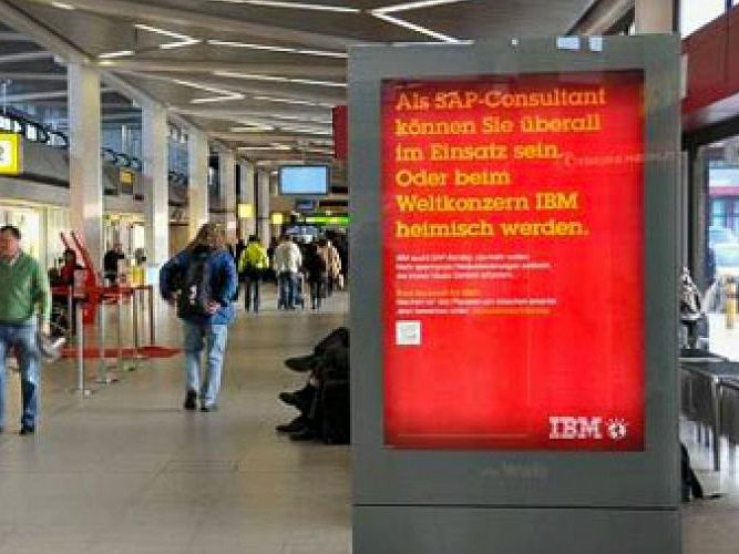 A City Light Poster at the check-in counter at Paris Airport. Many people are visible in the image. The poster advertising is related to IBM's personnel search.