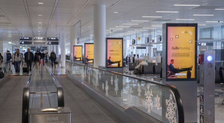 At Munich Airport, next to an escalator, four digital City Light posters can be seen. People are surrounding them. The digital screens display an image promotion for 'Gomeetings.com"