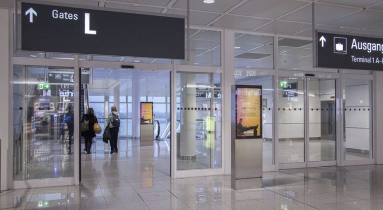 Through a passageway towards Gate L at Munich Airport, two digital City Light posters can be seen. One is in front of a door, while the other is behind the door next to an escalator. The digital advertising showcases an image promotion for 'Gomeetings.com'