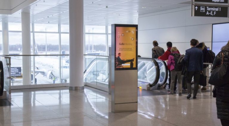 In the center of the image, next to an escalator, there is a digital City Light poster. It is located at Munich Airport, and many people are visible descending. The poster displays an image promotion for 'Gomeetings.com'