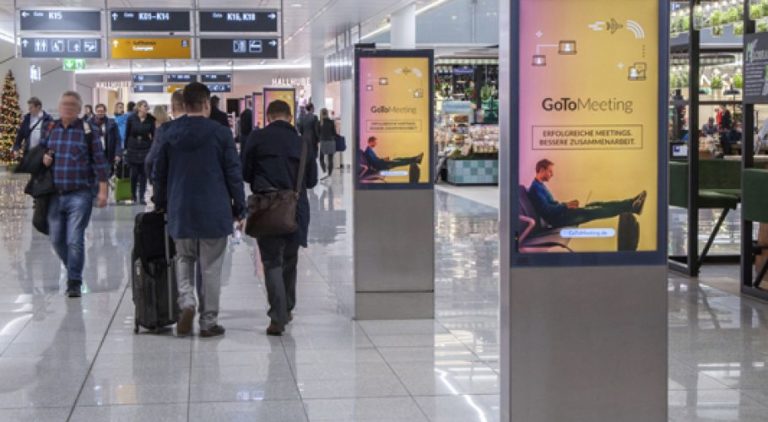At Munich Airport, two digital City Light posters are visible on the right side of the image. Many people are passing by. The advertising features an image promotion for 'Gomeetings.com'