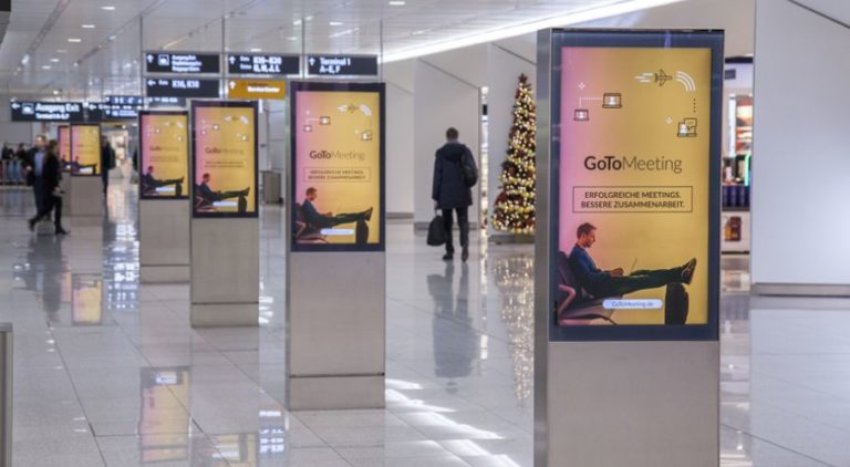 There are six digital City Light posters at Munich Airport, standing in a row. People are walking around them. The digital screens display an image promotion for 'Gomeetings.com'