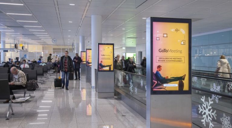 At Munich Airport, four digital City Light posters can be seen in a row next to an escalator. Seating areas are on the left side of the image, and several people are visible. The digital advertising showcases an image promotion for 'Gomeetings.com'