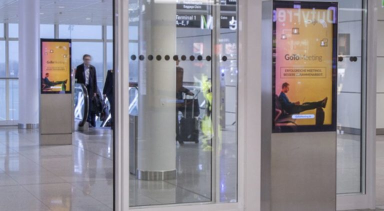 Two digital City Light posters are visible in Munich Airport. One is in the background on the left, and the other is in the foreground, right next to a passageway. The digital advertisement features an image promotion for 'Gotomeetings.com'