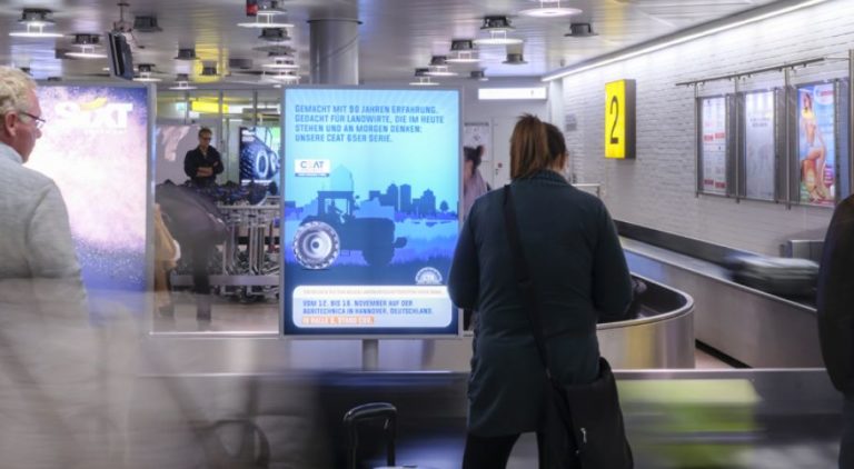 At Hannover Airport, a lightbox is visible at the baggage claim. Two people are standing in front of it. It displays an advertisement for CEAT.