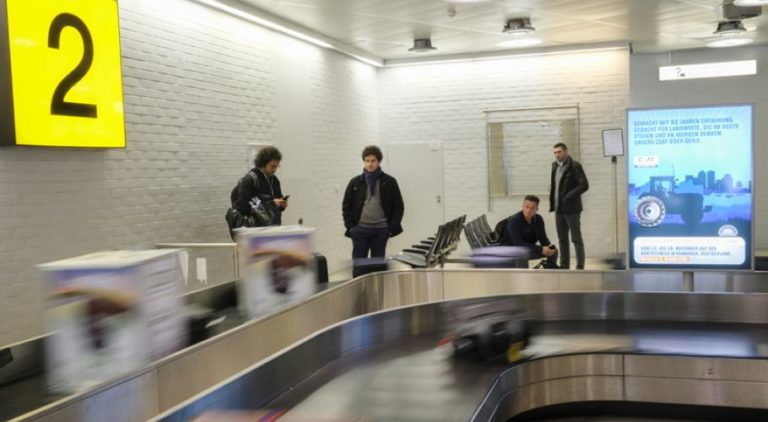 Passengers are waiting at the baggage claim for their luggage. Outdoor advertising is visible in the background.