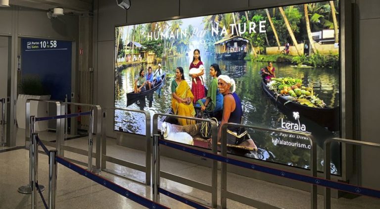 In the foreground, there is a lightbox at Paris Airport. The poster advertising displays tourism promotion from Kerala Tourism.