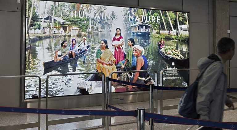 A large lightbox at Paris Airport, with a person passing by on the right side. The tourism poster advertising is from Kerala Tourism.