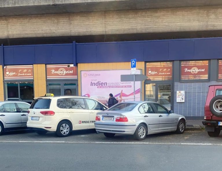 On a parking lot in Frankfurt, there is a large billboard behind the cars. It is attached to the wall of a bakery. It displays image advertising for the airline AirVistara.