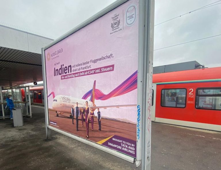 In the center of the image, there is a large billboard standing on a train platform in Frankfurt. On the billboard advertising, you can see the image advertising of the airline AirVistara.