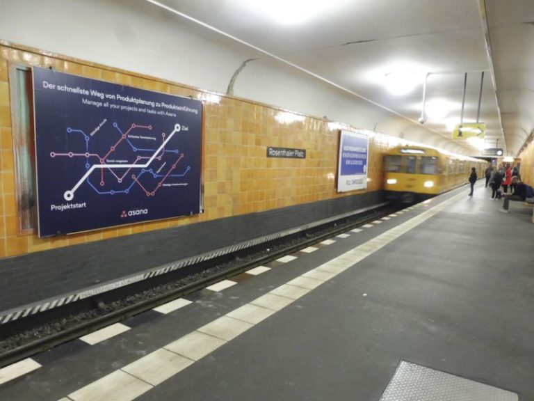 To the left in the foreground is a large advertising space. The large advertising space is at the 'Rosenthaler Platz' subway station in Berlin. In the background, a subway train is entering. The advertisement promotes a time management platform and is by the software company Asana.