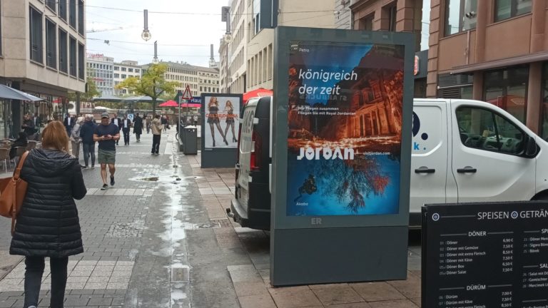In a pedestrian zone in Frankfurt am Main, people are walking and in the foreground on the right, there is a City Light poster. On the billboard advertisement, you can see tourism advertising for Jordan.