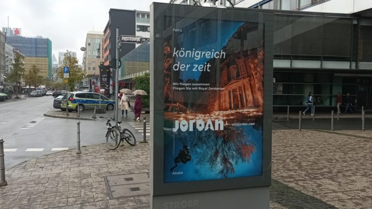 At an intersection in Frankfurt am Main, there is a City Light poster on the sidewalk. The billboard advertisement displays tourism advertising for Jordan.