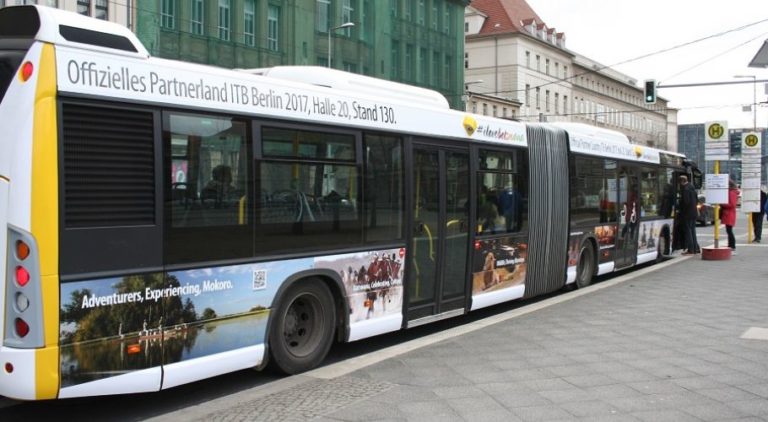 In the city center of Berlin, a bus with a Full Cover advertising motif of Botswana is standing at a bus stop where people board at the front.
