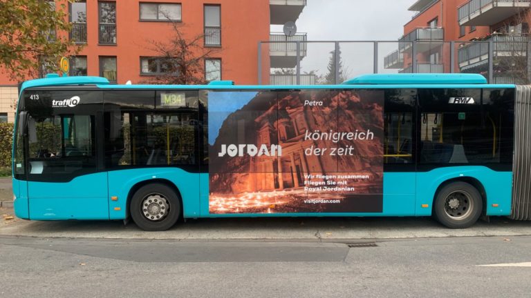 On a street in Frankfurt am Main, viewed from the front, a bus stop is visible. In the background, two residential blocks are recognizable. A bus with a side panel design is at the stop, and the design extends beyond the windows. The billboard advertising displays tourism promotion for Jordan.