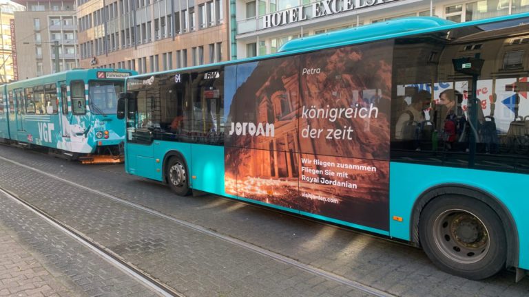 In downtown Frankfurt, viewed from the side, there is a bus behind a tram. The bus has a poster with tourism advertising for Jordan, while buildings are recognizable in the background.