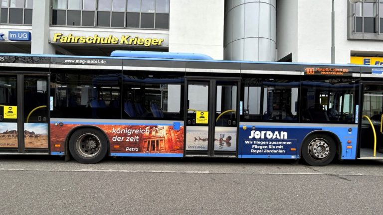 In Munich, on a street, a bus with a noticeable side panel design displaying tourism advertising for Jordan is driving.