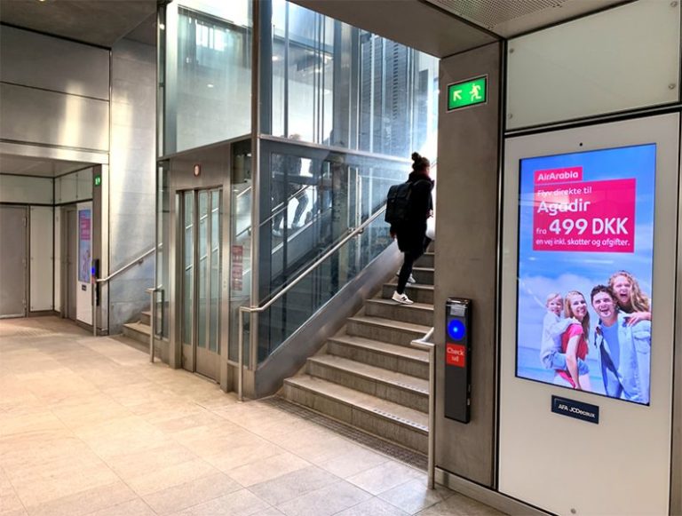In Copenhagen in the subway area, in the middle, an elevator is visible, flanked by stairs with a person walking up. Directly next to both stairs is the digital City Light poster with AirArabia advertising.