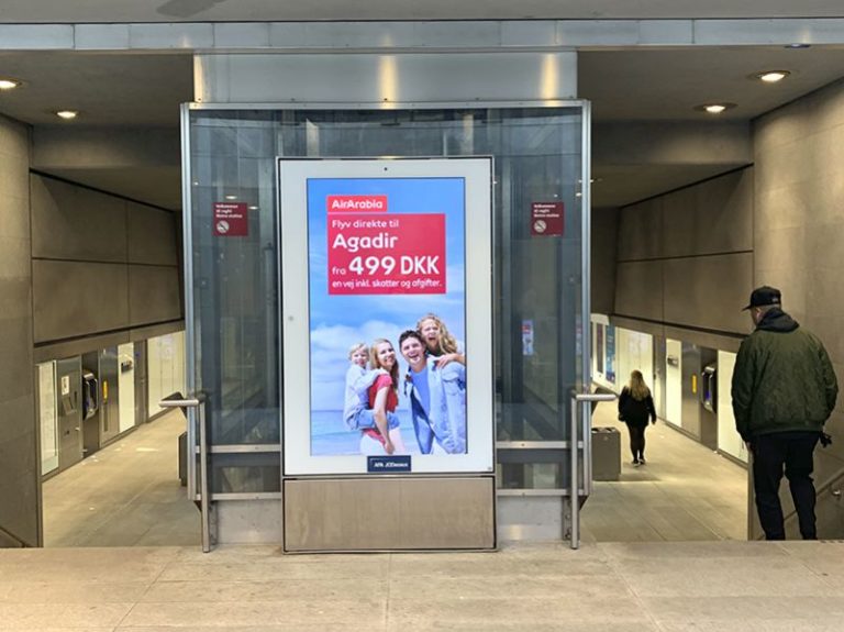 A digital City Light poster from AirArabia is visible in the center of the foreground behind an elevator in the subway area. Stairs go down on the left and right, and two pedestrians are walking down the stairs on the right.