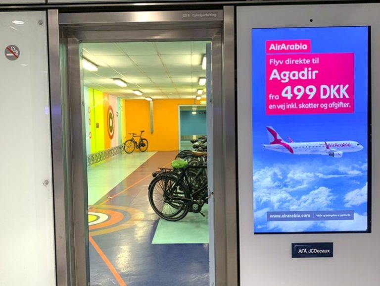 On the right side in the foreground, the digital City Light poster from AirArabia is visible. On the left side of the image, there is a glass entrance door leading to bicycle parking spaces.