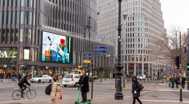 The image shows the shopping street Kurfürstendamm. On the left side of the image, a glass building is visible, with a large digital screen displaying Beluga Vodka advertising. In front of it, there is a street intersection with various participants in traffic.