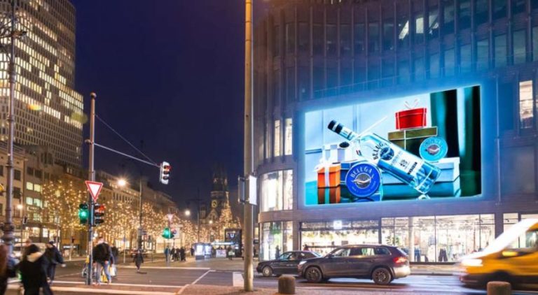 On the night-time shopping street Kurfürstendamm, on the left, a glass building is visible. On it, a large digital screen with Beluga Vodka advertising is displayed. At the intersection, pedestrians are crossing the street.