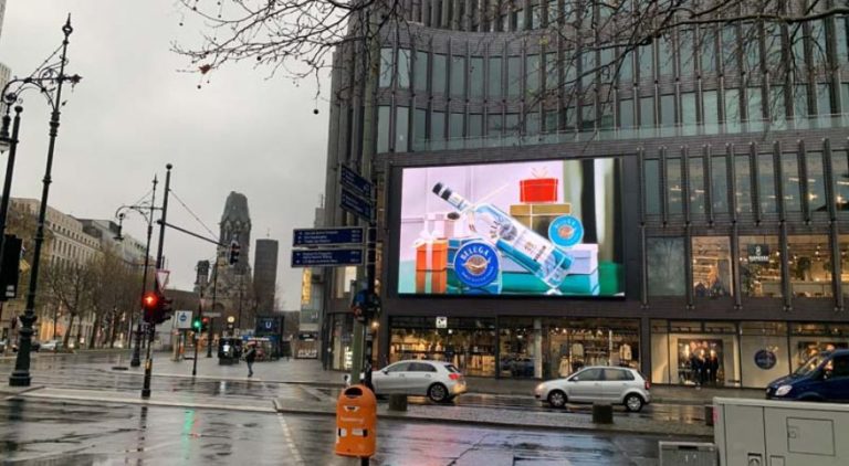 On the shopping street Kurfürstendamm, a glass building is visible. On it, there is a large digital screen displaying advertising for Beluga Vodka. On the left side of the image, in the background, you can see the Kaiser-Wilhelm Memorial Church.
