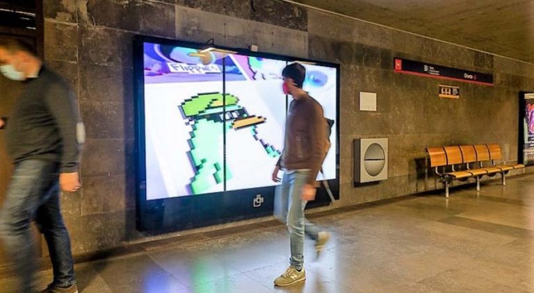 In the subway area of the Metro in Lisbon, a digital screen on the wall is visible. In the center of the image, a person is looking directly at the advertising from Flippies.art on the screen, while another person is passing from right to left. In the background on the right side, benches are visible.