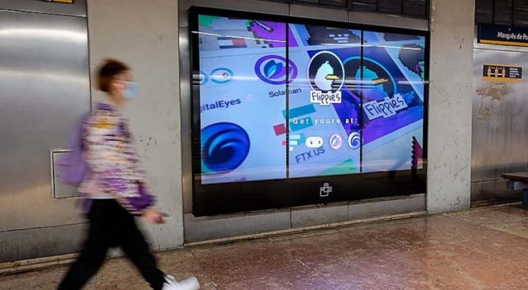 In Lisbon, in the subway area of the Metro, a digital screen is visible on a wall. On the left side of the image, a woman is walking, while in the background, the digital screen with advertising from the company Flippies.art is recognizable.