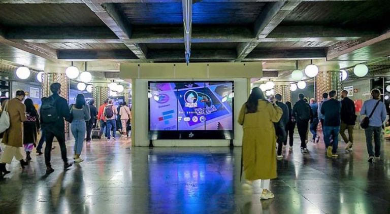 In the subway area of the Metro in Lisbon, there is a digital screen with advertising from Flippies.art, which is centrally visible in the background. People are moving on both sides of it.