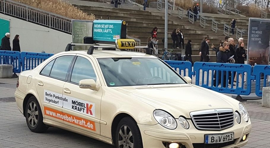 a taxi with a roof-top-ad, waiting in front of a trade fair. Trade fair visitors are passing by in the background.