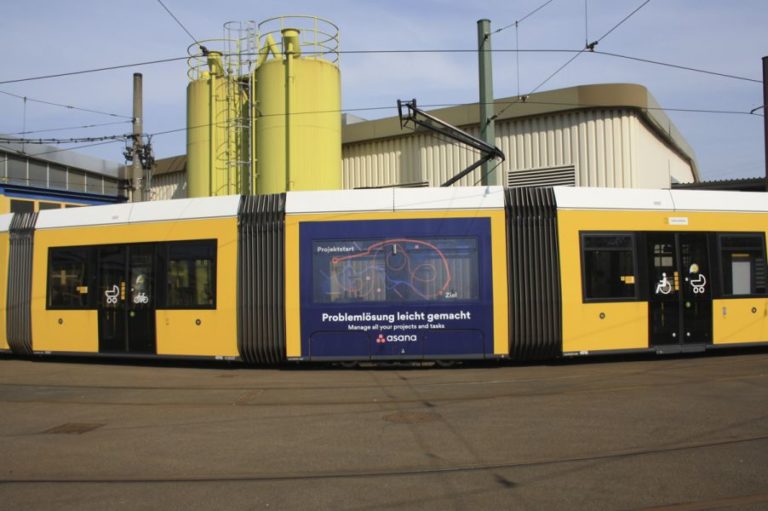 In an industrial area in Berlin, three carriages are seen head-on from a tram, featuring Traffic Board advertising from the company Asana. In the background, a factory building with two columns can be recognized.