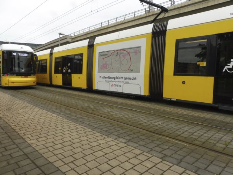 In Berlin, two trams are visible on the tracks. The entire tram on the right side is adorned with Traffic Board advertising from the Asana software company. On the left side, another tram is visible head-on.