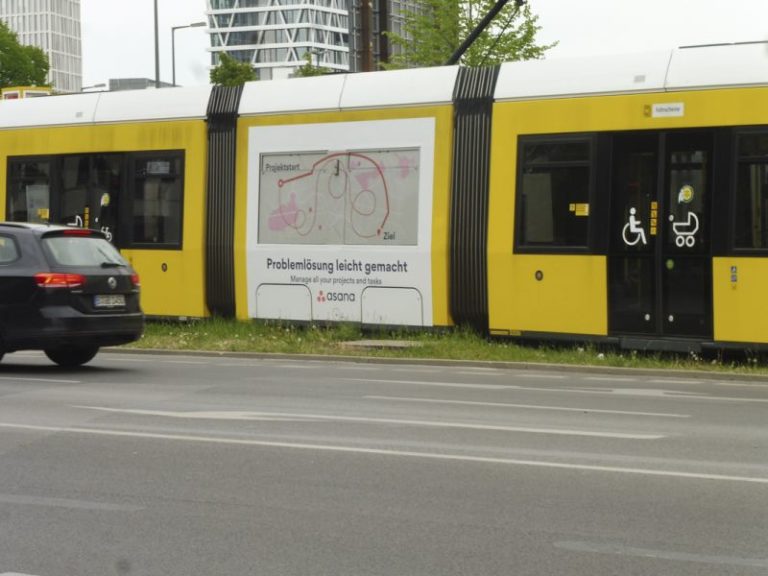 A tram is traveling in Berlin. Three carriages adorned with Traffic Board advertising from the software company Asana are visible. In front of the tram, there is a street with a car, and in the background, a building can be faintly recognized.