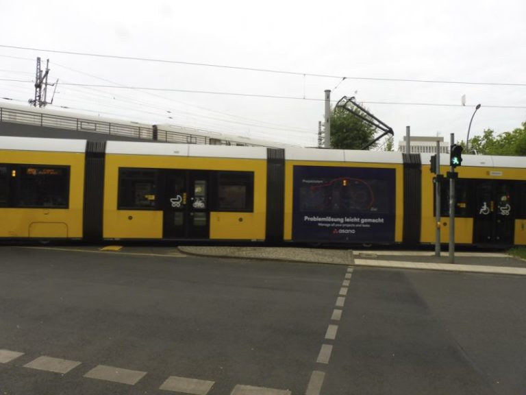 In Berlin, a tram with a view of four carriages decorated with Traffic Board advertising from the software company Asana turns left at an intersection. A street is visible in front of the tram.