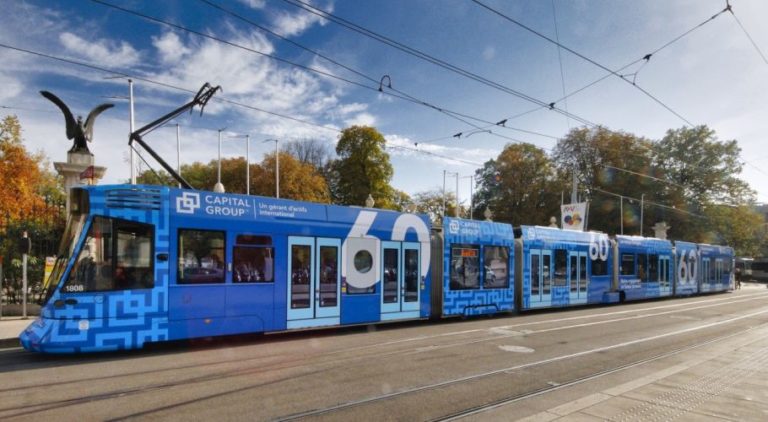 In Geneva, a tram is moving from right to left. In front of it, the street stretches, and behind it, there is a park. The tram is adorned with a blue Full Cover design promoting the Capital Group.