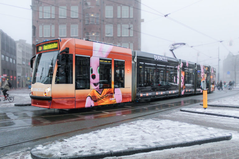 A tram adorned with a striking orange advertising motif is traveling through the snow-covered streets of Amsterdam