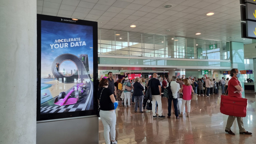 A person leans against a digital billboard at a crowded airport.