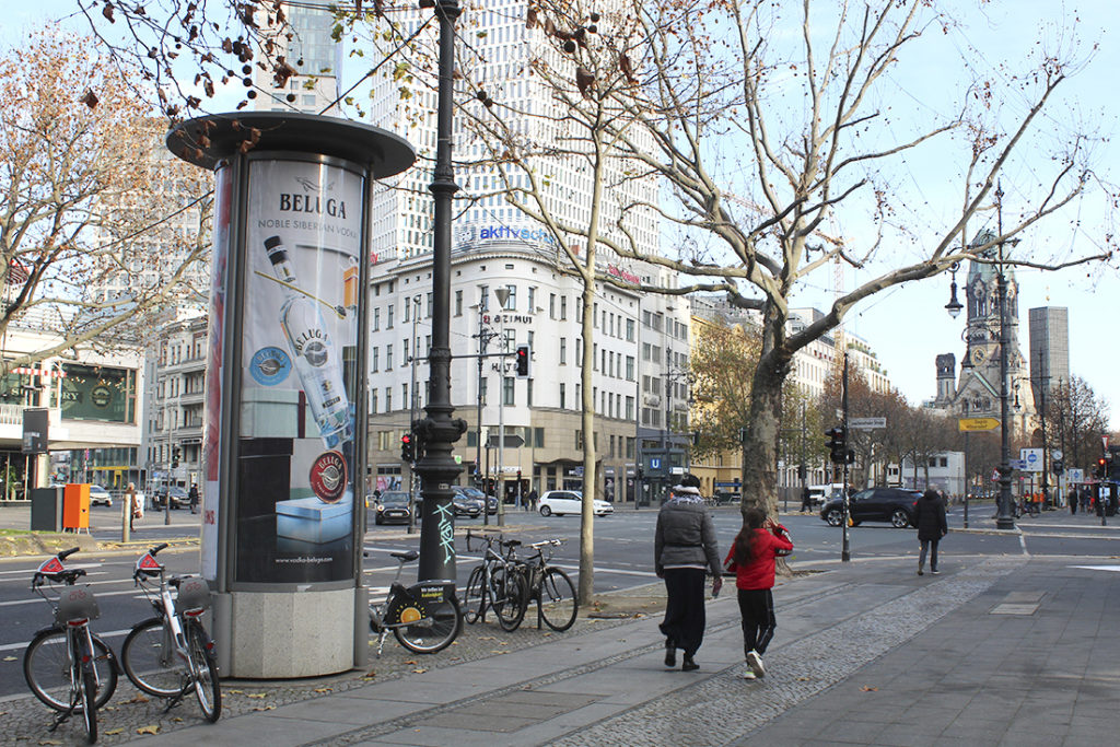 An advertising column featuring a Beluga advertisement stands in the middle of Berlin at a busy intersection.