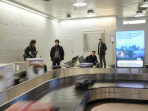 People are waiting at the baggage claim for their luggage. On the right, there is an advertising display.