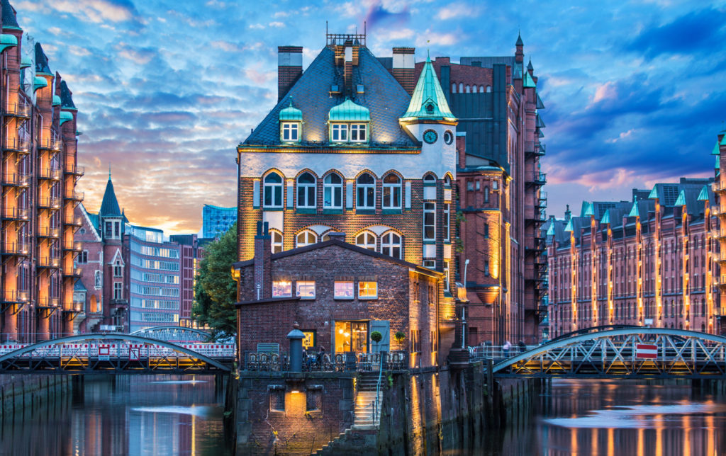 At nightfall, some buildings in Hamburg are visible in a river. They are connected by two bridges, while additional buildings extend in the surroundings
