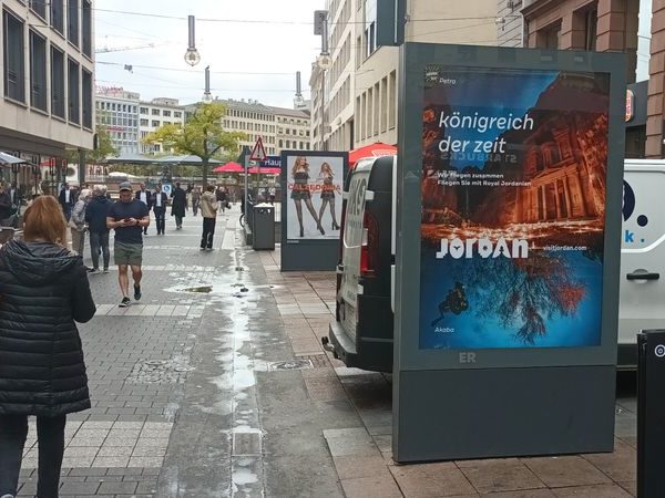 On a screen in a pedestrian zone, there is an advertising space for 'Jordan,' with many people strolling through the area.