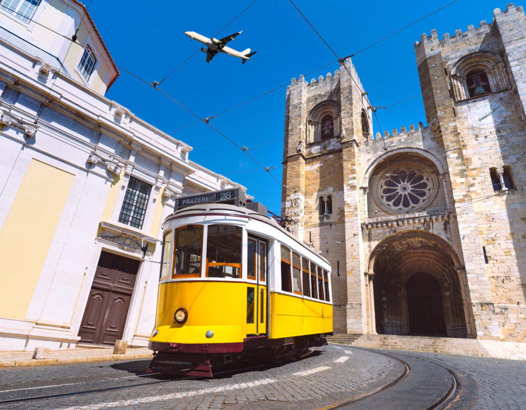A typical Lisbon tram is traveling through the old town. It is an older model in the classic yellow and white colors. In the background, historic buildings can be seen.