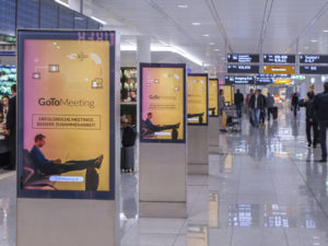 Numerous digital advertising displays are lined up in a row. They are positioned at an airport, promoting 'GoTo Meeting'.