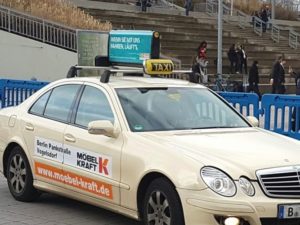 In the foreground, a taxi with a roof advertising sign from the company Sunnycars is visible. In the background, there are stair steps with people.
