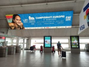 A big banner hangs at the airport in Rome, in the background are two digital screens.